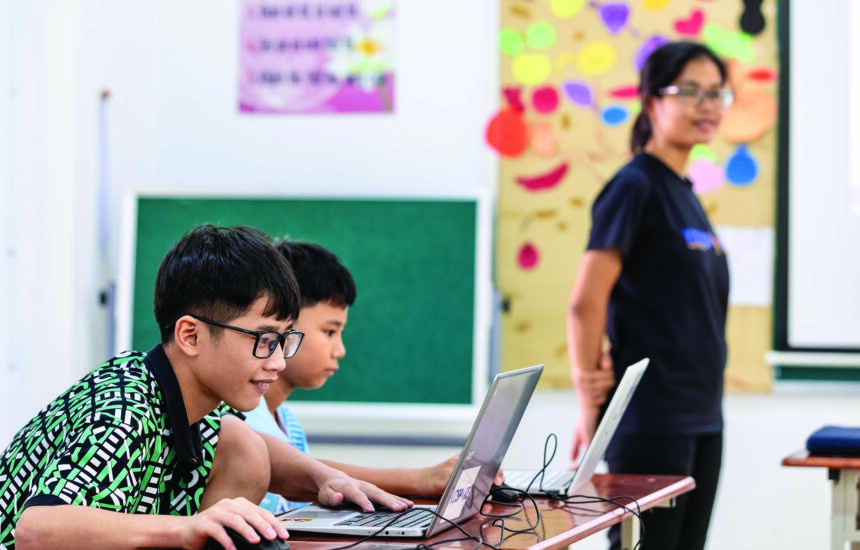 Two young students using laptops in a classroom, with a teacher students near them.