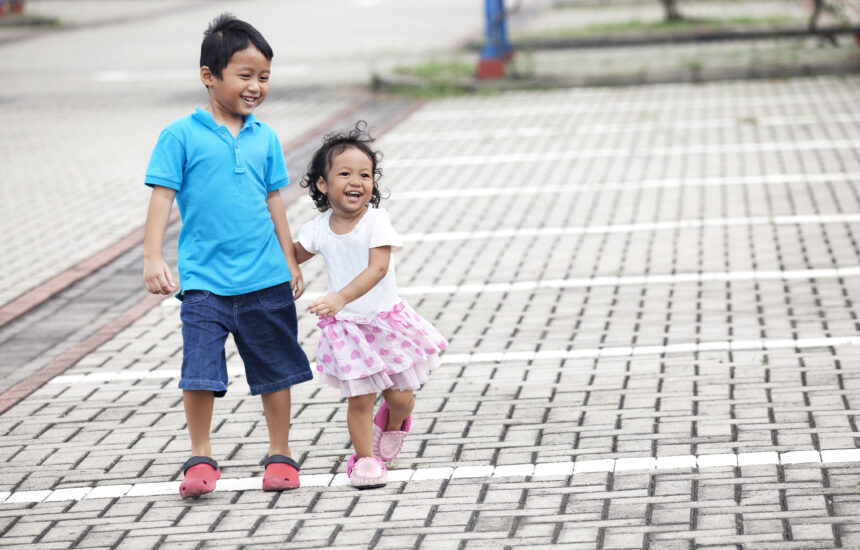 A photo of a young boy wearing a blue shirt and blue shorts holding hands with a younger girl wearing a white shirt and pink skirt. Both are smiling