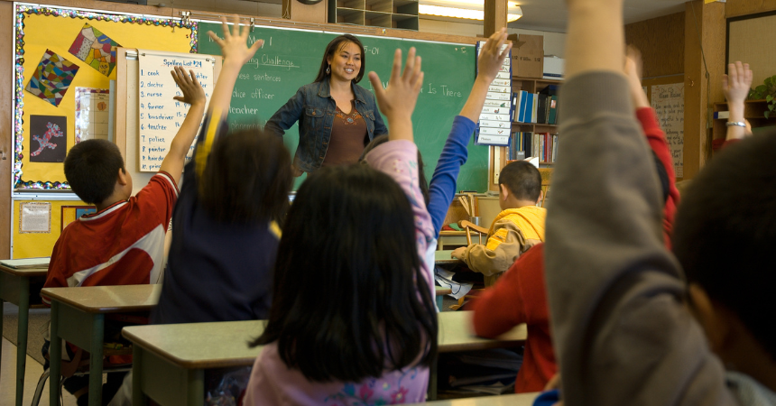 A teacher standing in front of a chalkboard, with students raising their hands in front of her.