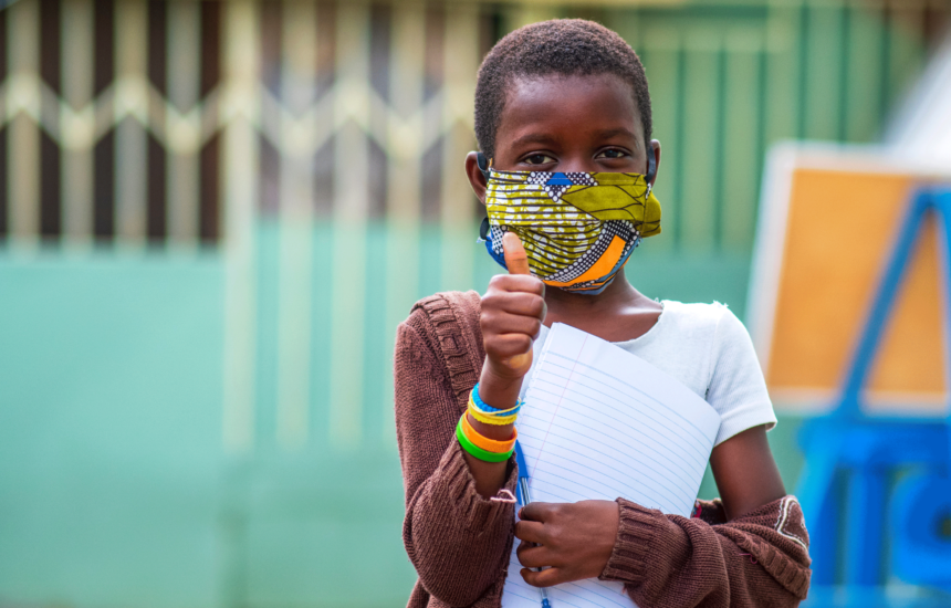 A child wears a face mask and holds a paper/pen and is giving a thumbs up