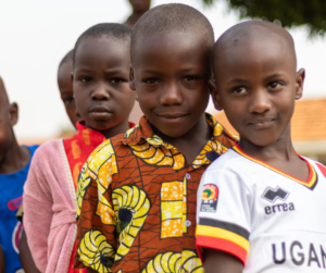 Group of young children standing close together outdoors. They have dark skin, brown eyes, and shaved heads and are wearing brightly colored clothing. They are smiling and look happy.