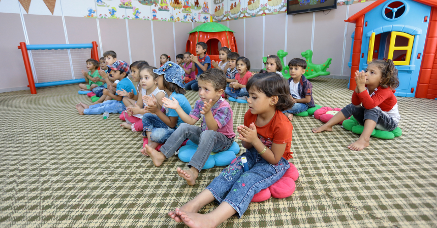 Young children who are refugees sitting on the floor of a school, repeating a song while clapping hands